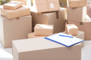 Clipboard, cardboard box and blurred stacked parcels on background, indoors